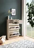 A white shoe cabinet with multiple shelves and drawers is shown in a room with a dark green wall and a large window. A rug with a geometric pattern is on the floor in front of the cabinet. Produktbild-Vorschau