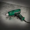 A green and black heat gun rests on a textured gray surface. Produktbild-Vorschau