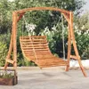 A wooden swing set with a curved wooden seat is set up outdoors on a paved patio, with lush green foliage and white flowers in the background. Podgląd zdjęcia produktu