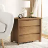 A wooden nightstand with three drawers is placed next to a white armchair, with a lamp, a clock, and a book on top. A window with curtains is in the background. Produktbild-Vorschau