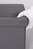 A hand wearing a white glove is pressing down on the corner of a gray upholstered storage ottoman. Produktbild-Vorschau