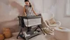 A woman is standing next to a baby crib in a nursery room. There is a rocking chair and an ottoman in the background. Produktbild-Vorschau