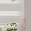 A close-up view of a white roller blind with a cassette, partially covering a window with greenery visible outside. Produktbild-Vorschau