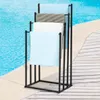 A black metal towel rack with four arms stands on a wooden deck next to a swimming pool. Produktbild-Vorschau