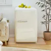 A cream-colored retro-style mini-fridge is placed on a light-colored mat on a wooden floor, next to a sofa and a potted plant. Lemons are arranged on top of the fridge. Produktbild-Vorschau