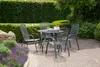 A round garden table with four chairs set on a paved patio, surrounded by lush green hedges and purple flowers. Produktbild-Vorschau