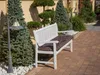 A white garden bench with a brown cushion sits on a brick patio, surrounded by evergreen trees and decorative stones. Náhled obrázku produktu