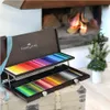 A wooden case of colored pencils is open, displaying rows of pencils, in front of a fireplace with a Christmas tree nearby. Podgląd zdjęcia produktu