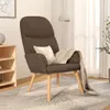 A brown fabric armchair with a beige throw blanket and pillow, placed on a patterned rug in a room with a wooden shelf and a window with sheer curtains. Náhľad obrázku produktu