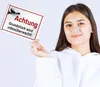 A young woman is holding a metal sign that says "Achtung! Grundstück wird videoüberwacht!" (Attention! Property is under video surveillance!). Produktbild-Vorschau