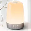 A bedside lamp with a digital clock display is shown on a table next to a vase with flowers. Produktbild-Vorschau