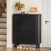 A black shoe cabinet with roll-up doors is placed in a hallway next to a staircase. Decorative items like flowers, books, and a handbag are on top of the cabinet. Several pairs of slippers are neatly arranged on the floor in front of the cabinet. Produktbild-Vorschau
