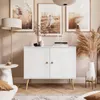 A white two-door cabinet with wooden legs and handles, decorated with vases and books, in a room with artwork and a floor lamp. Produktbild-Vorschau