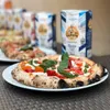 A close-up of a pizza on a white plate, with bags of flour in the background. Produktbild-Vorschau