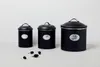 Three black storage containers of varying sizes are arranged on a white surface against a white background. Small dried seed pods are scattered in front of the containers. Produktbild-Vorschau