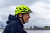 A woman wearing a yellow bicycle helmet smiles while looking to the right. The background is a cloudy sky and a body of water with some structures in the distance. Náhľad obrázku produktu