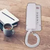 A white landline telephone is mounted on a wooden desk next to a notebook and a cup of coffee. Podgląd zdjęcia produktu