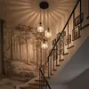 A staircase with a forest-themed wallpaper and a black metal railing adorned with framed pictures. Three pendant lights hang from the ceiling above the stairs. Náhled obrázku produktu