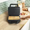 A waffle maker with two waffles inside, and a hand using tongs to remove a waffle. Podgląd zdjęcia produktu