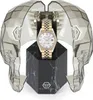 A gold watch with diamond accents is displayed on a black marble stand, inside a transparent, faceted skull-shaped case. Podgląd zdjęcia produktu