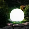 A glowing white sphere light sits on grass in a garden, surrounded by rocks and foliage. Produktbild-Vorschau