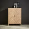 A wooden cabinet with a herringbone pattern on the doors, placed against a dark wall. Produktbild-Vorschau