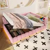 A pink upholstered bed with its storage compartment open, revealing folded items. The bed is made up with bedding and pillows, and is placed on a rug with a cross pattern. The room has a modern, minimalist aesthetic with decorative shelves and lighting.