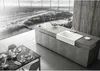 A modern kitchen with a large window overlooking a city view, featuring a kitchen island with a sink and a dining table set for breakfast. Produktbild-Vorschau