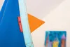A close-up of a blue fabric product with an orange triangular accent and two red tags. A blurred painting of houses is visible in the background. Náhled obrázku produktu