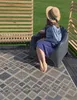 A person sits in a gray chair on a patterned rug outdoors, with a wooden fence and green grass in the background. Produktbild-Vorschau
