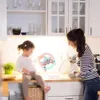 Rosa und hellblaues Spielzeuglenkrad, gehalten von einem Kind, das auf einer Küchenarbeitsplatte sitzt, neben einer Frau, die Geschirr spült Náhľad obrázku produktu