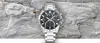 A silver chronograph watch with a black dial is displayed centrally against a textured gray rock background. Voir les images du produit