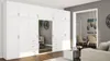 A large white modular wardrobe with mirrored doors is shown in a modern living room setting, with a glimpse into a dining area through an open doorway. Podgląd zdjęcia produktu