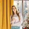 A young woman holding a mug stands by a window with a Christmas tree and snowy scenery outside. Produktbild-Vorschau