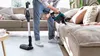 A person is using a cordless vacuum cleaner to clean a sofa in a living room. Produktbild-Vorschau