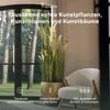 A living room scene with a large artificial plant in a golden pot next to a white armchair, with a large window showing trees in the background. Produktbild-Vorschau