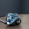 A blue bagless vacuum cleaner is shown in a room with dark gray walls and a light gray floor. A black power cord snakes across the floor to an electrical outlet. Produktbild-Vorschau