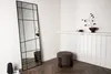 A tall, rectangular mirror with a black grid frame stands in a corner next to a small, round, brown ottoman. A decorative branch with dark buds sits on a white shelf above the ottoman. Podgląd zdjęcia produktu