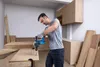 A man is using a cordless drill to drill into a piece of wood in a workshop setting. Produktbild-Vorschau