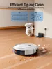 A robot vacuum cleaner is cleaning a wooden floor, with a small white dog in the background. Podgląd zdjęcia produktu