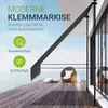 A modern clamp awning is shown extended over a wooden deck with a view of the ocean. The awning is gray and supported by black poles. There is a wicker chair with cushions on the deck. Náhľad obrázku produktu