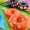 A close-up of several decorated donuts with candy eyes, arranged on colorful paper plates on a green surface, with confetti scattered around. Náhled obrázku produktu
