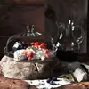 A cheese platter with grapes and tomatoes under a glass dome, served on a rustic wooden stand, with a carafe of red wine in the background. Náhľad obrázku produktu