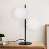 A modern black table lamp with two white glass globes sits on a wooden surface next to a book and a small plant. Náhľad obrázku produktu