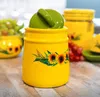 A yellow ceramic jar with a sunflower design and a green lid sits on a wooden table, with other jars and grapes in the background. Produktbild-Vorschau