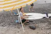 A man is sitting on a beach under a striped umbrella, holding a surfboard. There are other surfboards and a portable speaker on the beach. Produktbild-Vorschau