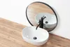 A white oval countertop sink with a black faucet is shown on a wooden surface, with a round mirror reflecting the sink and faucet in the background. Anteprima dell’immagine prodotto