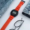 A close-up shot of a bright orange digital watch with a black face, placed next to a pair of glasses and a folded denim jacket. Anteprima dell’immagine prodotto