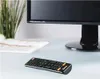 A remote control is placed on a desk in front of a computer monitor. Voir les images du produit