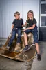 Two children are sitting in a large excavator bucket in front of a building. Produktbild-Vorschau